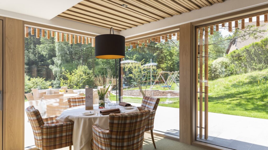 Heller Essbereich mit gedecktem Tisch und karierten Stühlen, neben großen Glasfenstern mit Blick auf einen sonnigen Garten mit Spielplatz im nachhaltigen Hotel Weihrerhof in Südtirol, der eine ruhige Atmosphäre schafft.