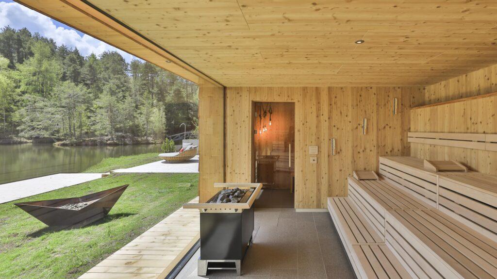 Moderne Sauna mit Holzinterieur und Blick auf einen ruhigen See und üppige Bäume im nachhaltigen Wellnesshotel Weihrerhof in Südtirol. Draußen befinden sich eine Feuerstelle und Liegestühle, die eine ruhige, entspannte Atmosphäre schaffen.