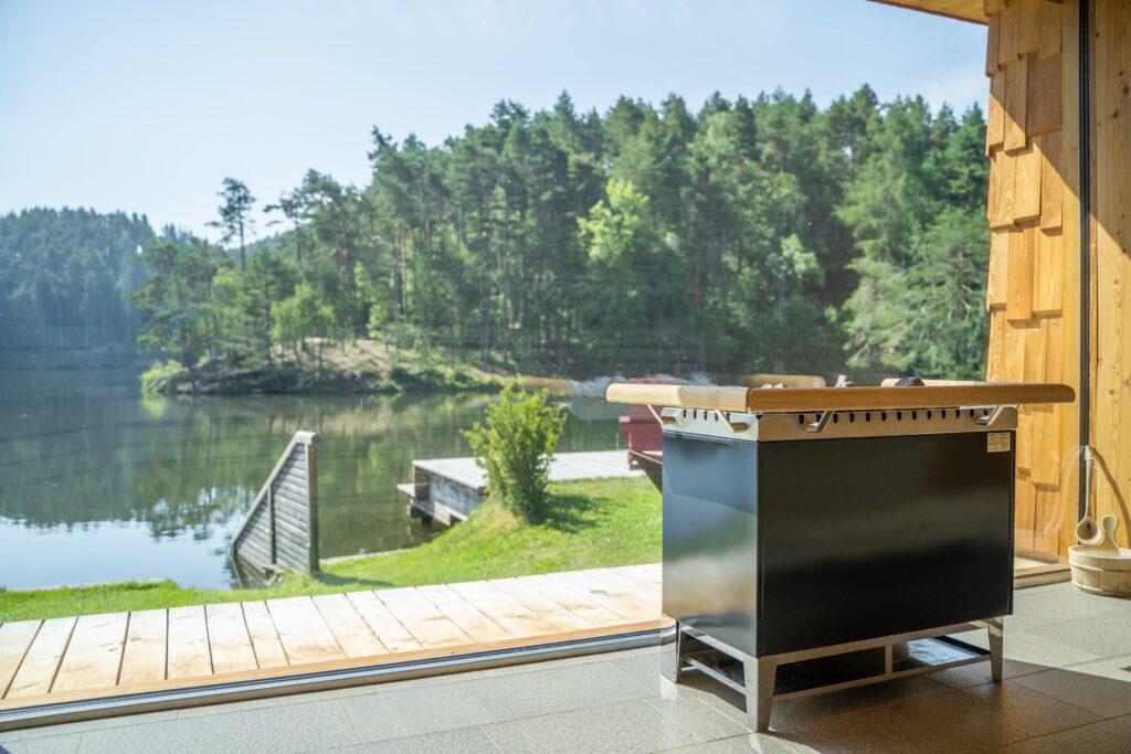 Ein moderner Holzofen in der einzigartigen Bootshaussauna des Südtiroler Hotels Weihrerhof, mit Blick auf einen ruhigen See und dichten Wald durch ein großes Fenster. Die Szenerie ist friedlich und einladend.