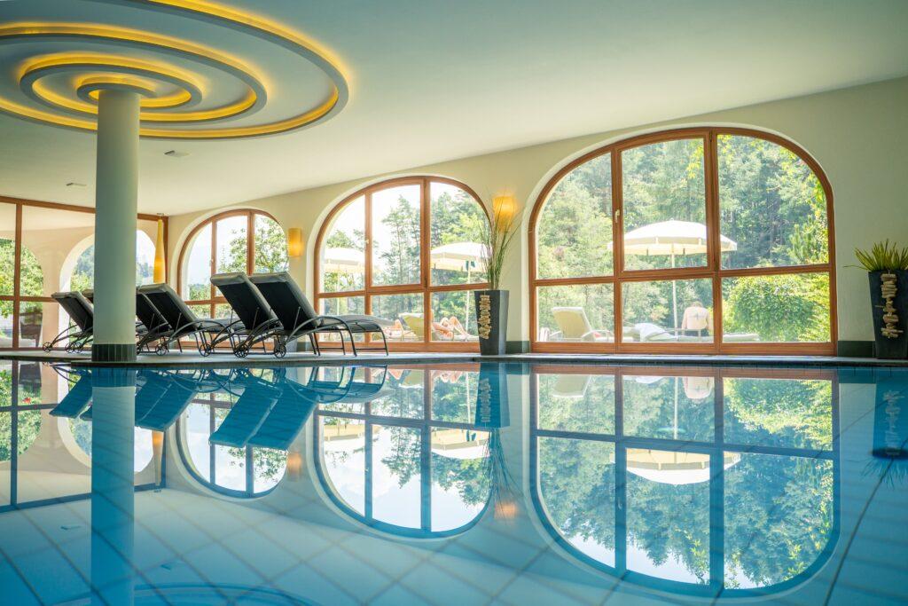 Luxuriöser Innenpoolbereich mit Liegestühlen, geschwungenen Fenstern und ruhigen Reflexionen auf dem Wasser. Draußen sorgt üppiges Grün für eine entspannte Atmosphäre.