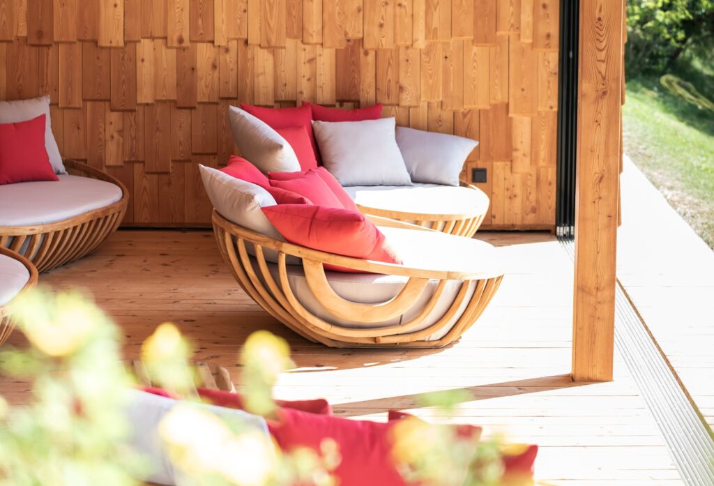 Gemütliche Holzterrasse mit geschwungenen Liegestühlen, roten und grauen Kissen, vor einer holzgetäfelten Wand und einem sonnendurchfluteten Außenbereich.