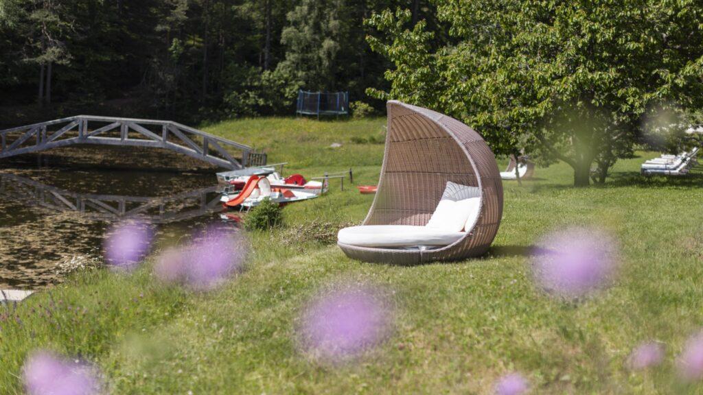 Korb-Liegestuhl mit Kissen auf einer Rasenfläche an einem ruhigen See im Hotel Weihrerhof in Südtirol. Eine Holzbrücke, Ruderboote und violette Blumen schaffen eine ruhige, entspannende Atmosphäre.