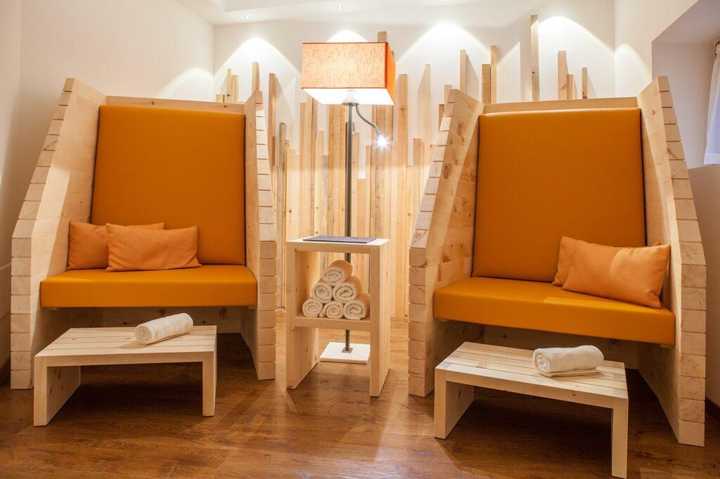Gemütliche Lounge im SeaSpa des Hotel Weihrerhof in Südtirol mit zwei orange gepolsterten Holzsitzen, Beistelltischen mit gerollten Handtüchern. Eine warme Lampe wirft sanftes Licht und schafft eine ruhige, einladende Atmosphäre.
