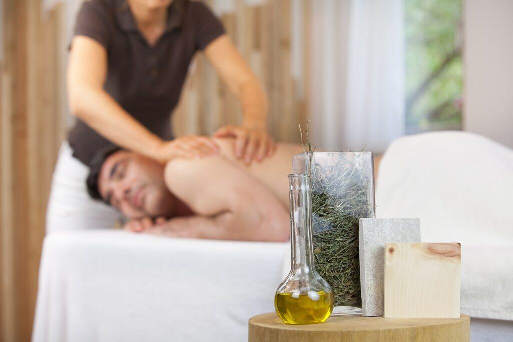 Eine Person erhält eine Rückenmassage in einer ruhigen Spa-Umgebung. Im Vordergrund sind Massageöl und Kräuter zu sehen, die eine ruhige, erholsame Atmosphäre schaffen.
