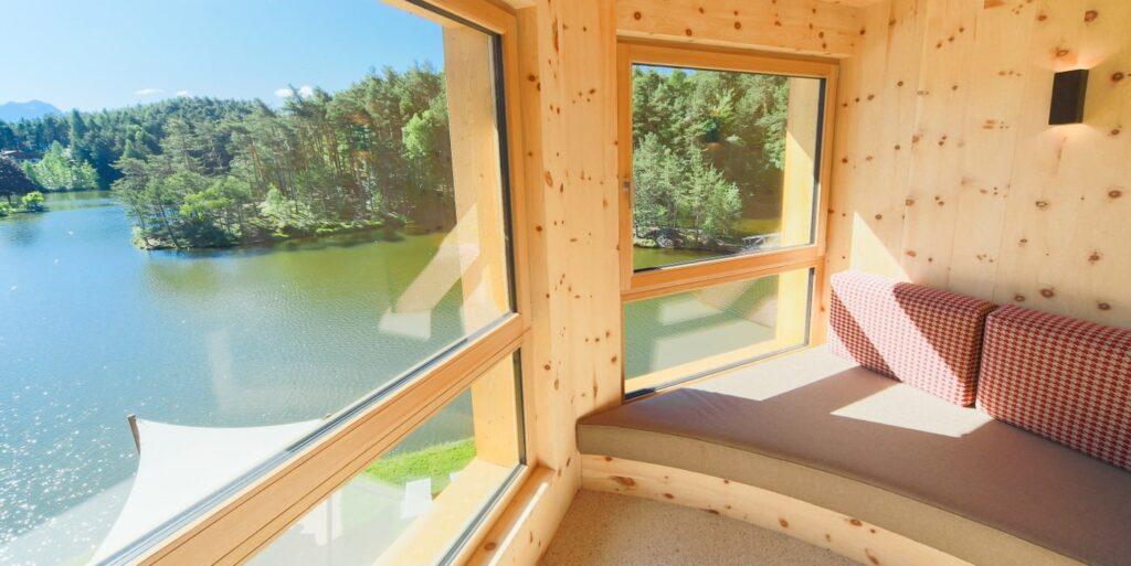Gemütliches Hotelzimmer aus Holz mit großen Fenstern und Blick auf den Wolfsgrubensee in Südtirol und den üppig grünen Wald. Eine geschwungene gepolsterte Bank mit gemusterten Kissen sorgt für zusätzliche Wärme. Hotel Weihrerhof.
