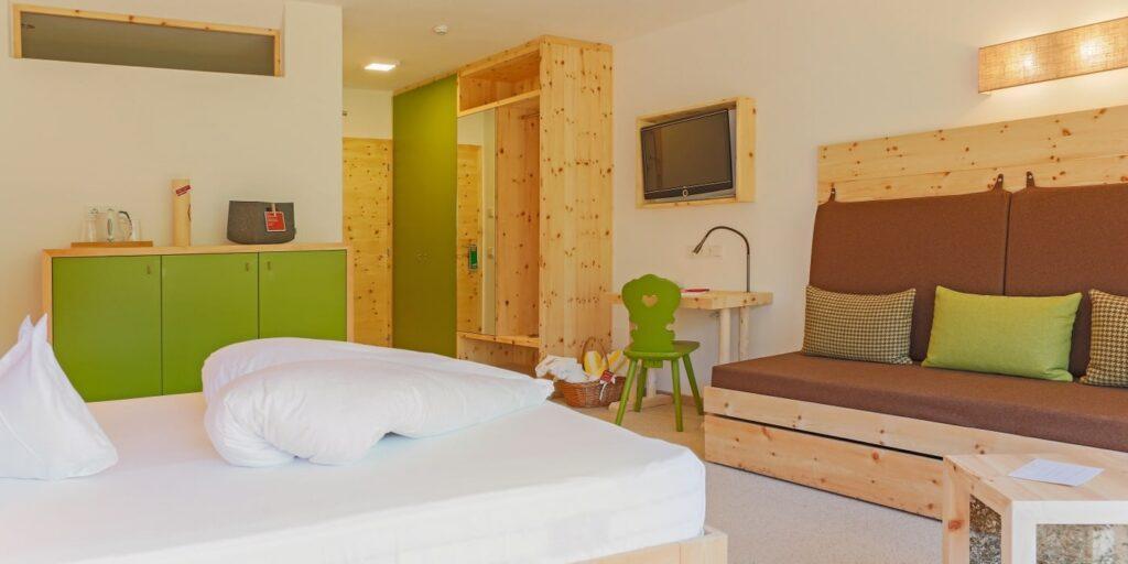 Modernes Hotelzimmer im nachhaltigen Hotel Weihrerhof in Südtirol mit hellen Holzmöbeln und grünen Akzenten. Mit ordentlich gemachtem Bett, gemütlicher Sitzecke, Wandfernseher und sanfter Beleuchtung.