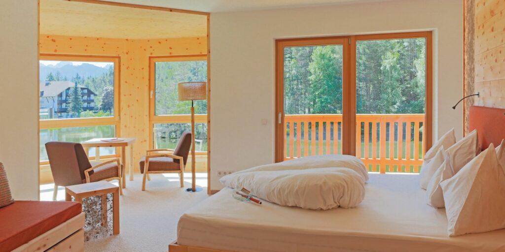 Helles, gemütliches Hotelzimmer im nachhaltigen Hotel Weihrerhof in Südtirol mit einem großen Bett vor Glastüren, die auf einen Balkon mit Blick auf den Wald führen. Der sonnendurchflutete Sitzbereich am Fenster sorgt für eine ruhige Atmosphäre.