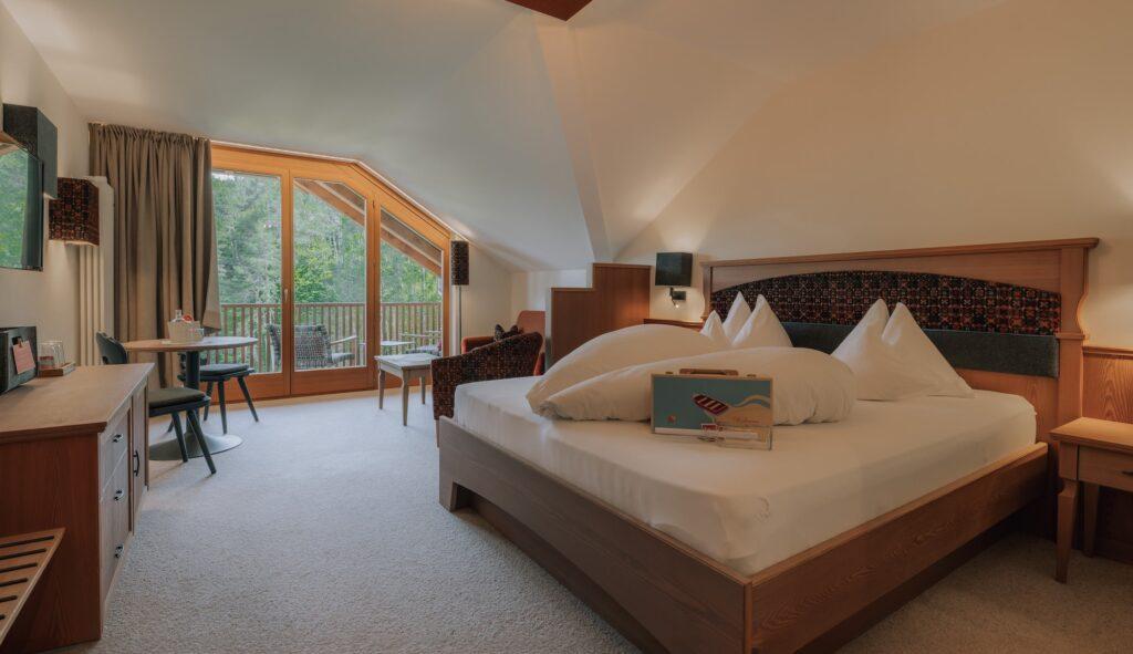Gemütliches Cradle-to-Cradle-Hotelzimmer im nachhaltigen Hotel Weihrerhof in Südtirol mit großem Bett, Holzmöbeln und Balkon mit Blick auf üppige Bäume. Sanfte Beleuchtung schafft eine warme, einladende Atmosphäre.
