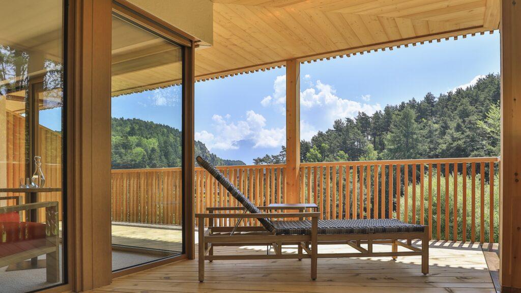 Moderner Holzbalkon mit einer einzelnen Chaiselongue, mit Blick auf einen üppig grünen Wald und einen blauen Himmel mit flauschigen Wolken. Die Szene ist ruhig und einladend.