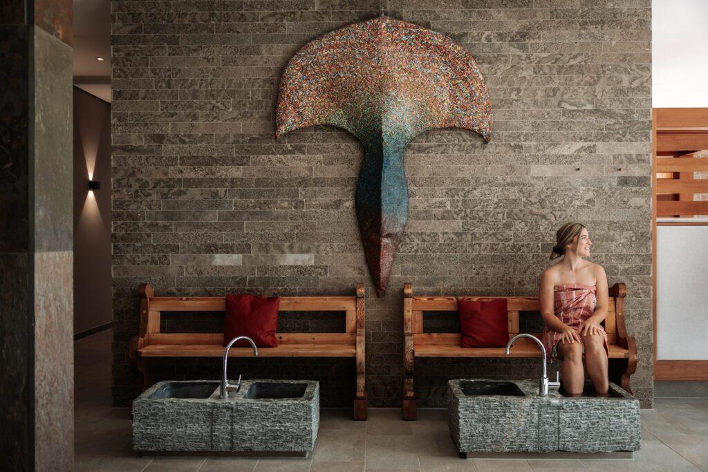 A woman wearing a towel sits on a wooden bench with red cushions next to stone washbasins in the sustainable Artemacur Spa at the Klosterhof wellness hotel in Bavaria. A colorful sculpture in the shape of a whale's tail hangs on the brick wall behind her.