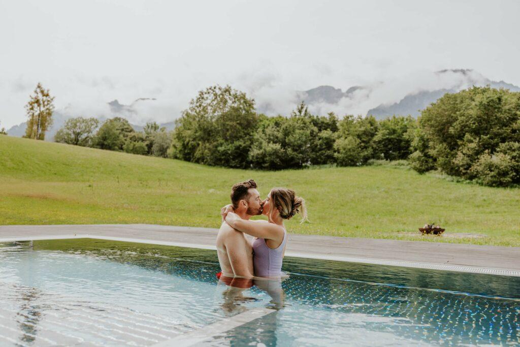 A couple embraces in the heated outdoor pool of the sustainable wellness hotel Klosterhof in Bavaria, surrounded by green hills and misty mountains. The scene appears tranquil and romantic, with a cloudy sky above.