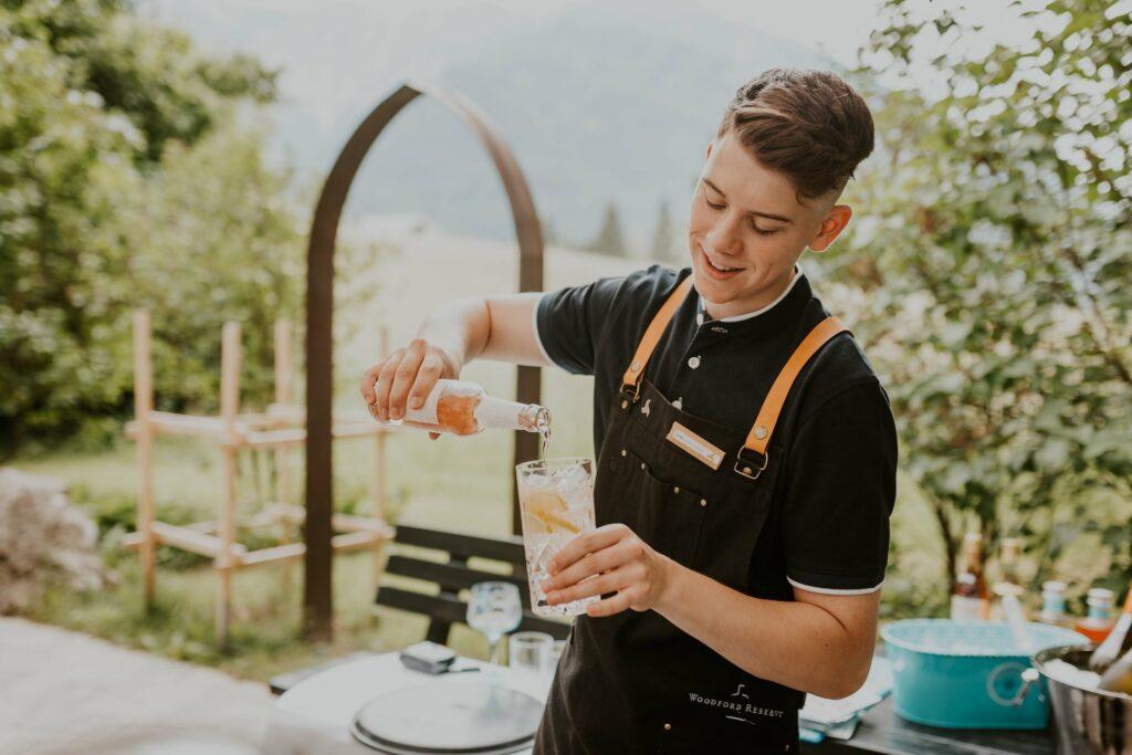 A young bartender smiles as he pours a drink into a glass outside. He is wearing a black apron with brown straps. The background is lush and green.