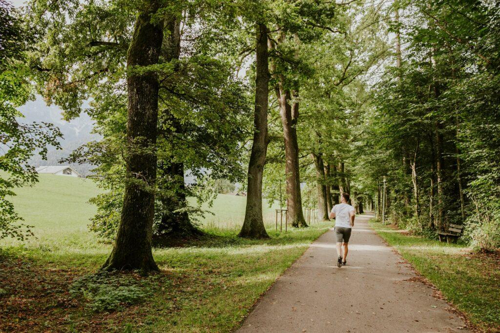 A man jogs along a tree-lined path, surrounded by lush greenery and wide fields under a cloudy sky, conveying a sense of tranquility and nature.