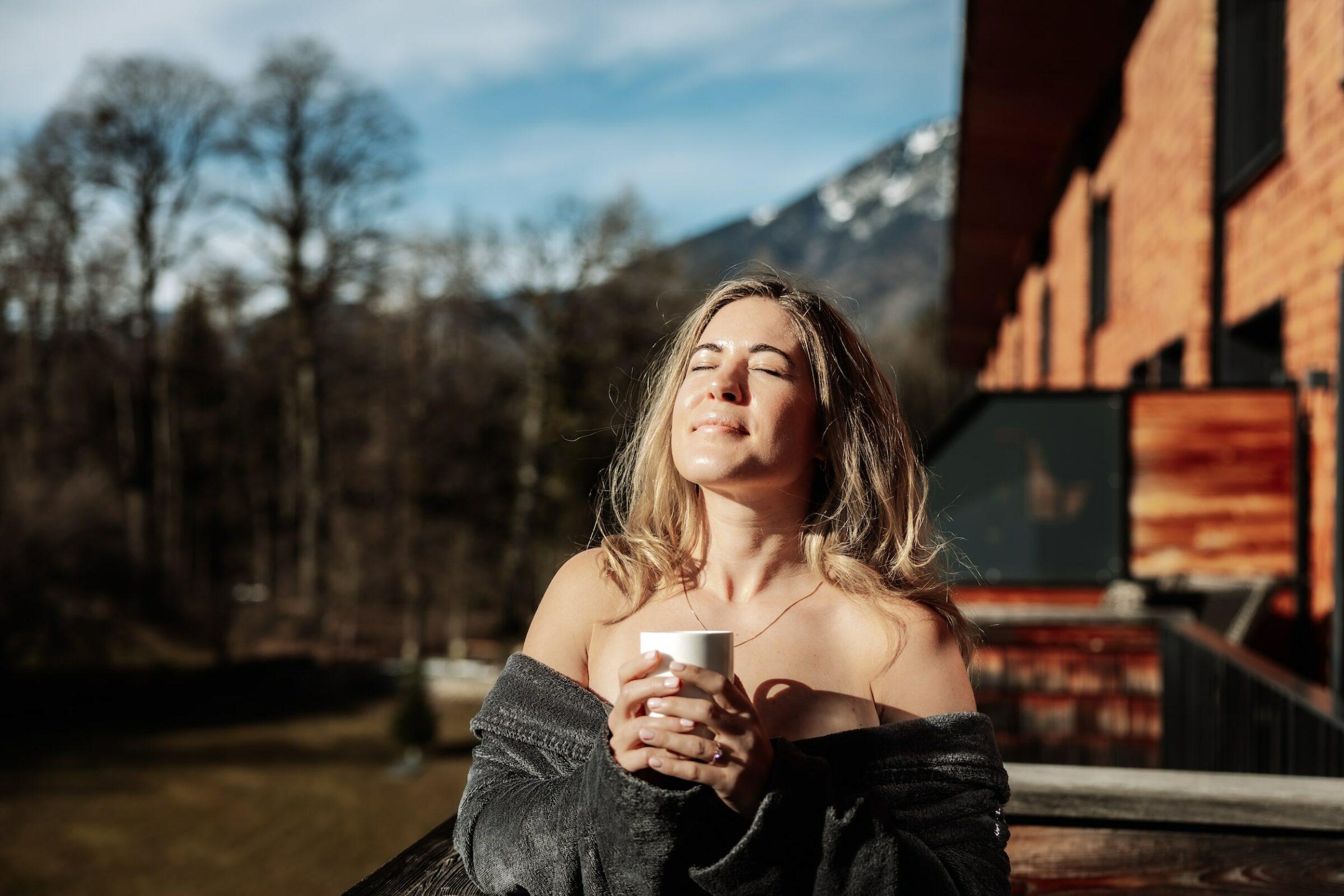 A woman enjoys the warm rays of sunshine on the balcony of the sustainable wellness hotel Klosterhof – Alpine Hideaway & Spa near Salzburg, holding a cup in her hand. She is wrapped in a cozy sweater, her eyes closed, looking serene. Snow-capped mountains can be seen in the background.