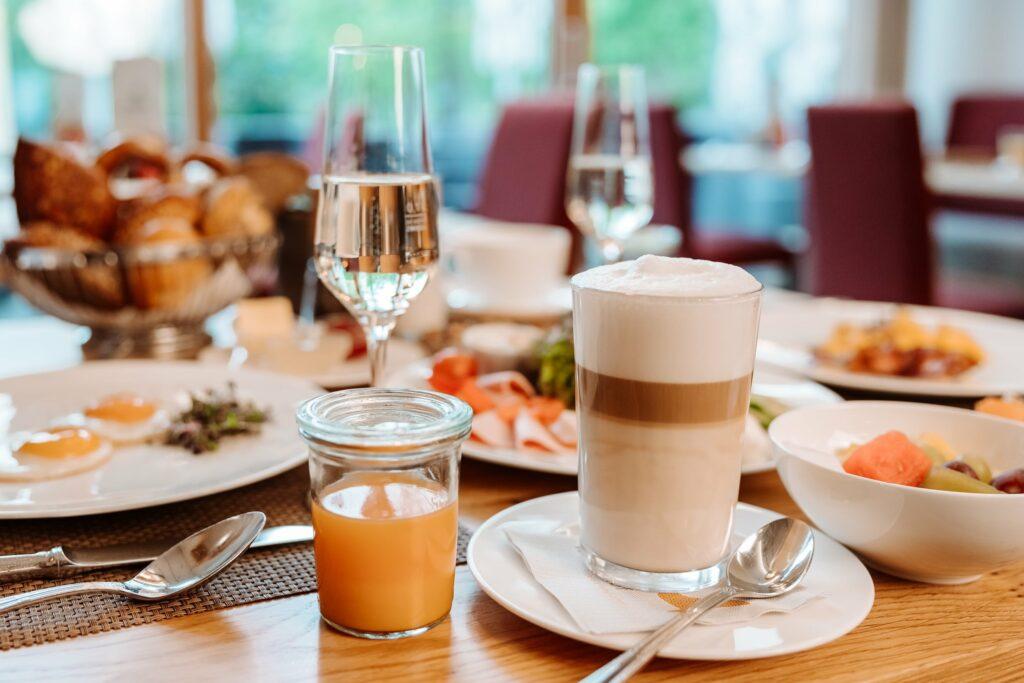 A cozy breakfast scene in the monastery courtyard – Alpine Hideaway & Spa in Bayerisch Gmain with cappuccino, orange juice, and sparkling wine on a table. Next to it are plates with eggs, fruit, and pastries, creating a relaxed, inviting atmosphere.