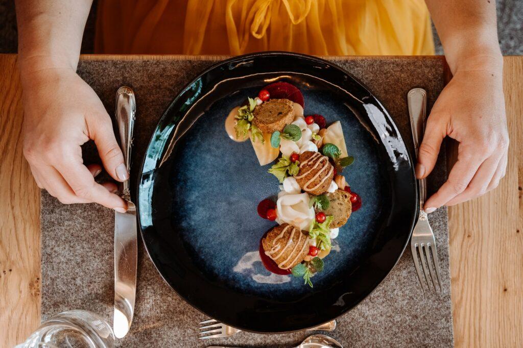 A person dressed in yellow sits at a wooden table holding cutlery next to a stylish black plate with an artfully arranged plant-based gourmet dish at the sustainable Hotel Klosterhof in Bavaria.