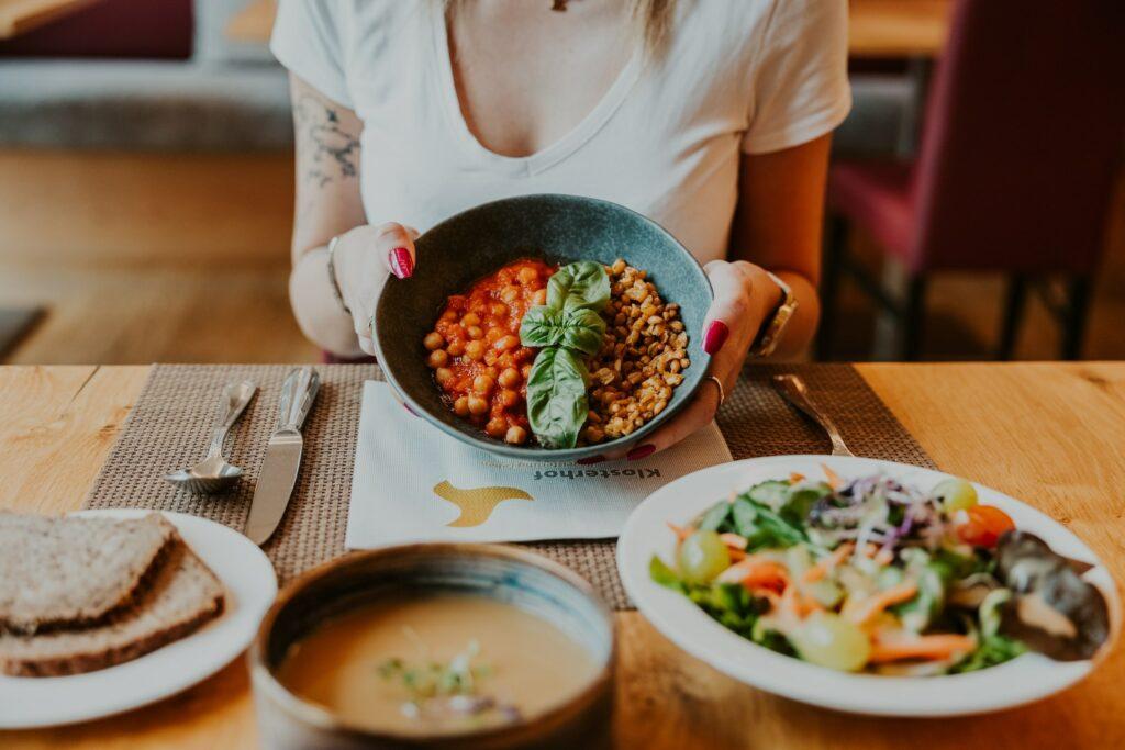 A woman in a white shirt holds a bowl of chickpeas and grains garnished with basil. Soup, salad, and bread are laid out on the table, creating a cozy lunchtime scene at the Wellnesshotel Klosterhof – Alpine Hideaway & Spa near Salzburg.