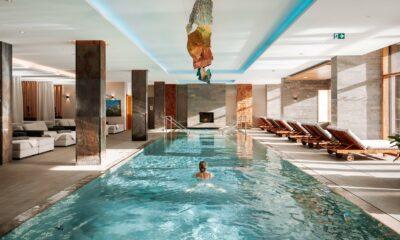 Innenpool des Wellnesshotels Klosterhof in Bayern mit einer Person, die unter einer modernen Kunstinstallation an der Decke schwimmt. Der sonnendurchflutete Raum lädt mit Liegestühlen und weichen Sofas zum Entspannen ein und schafft eine ruhige Atmosphäre.