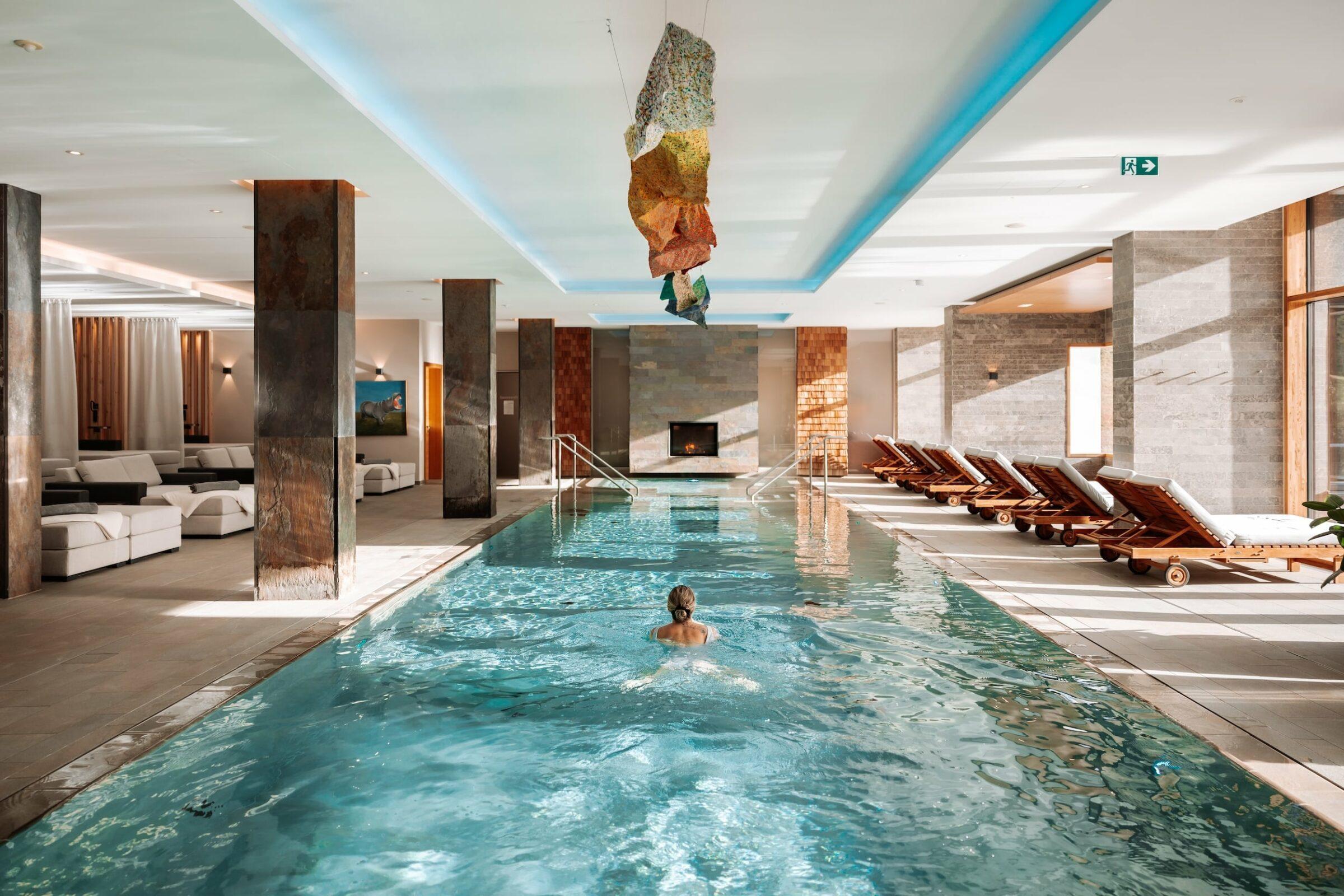 Indoor pool at the Klosterhof wellness hotel in Bavaria with a person swimming beneath a modern art installation on the ceiling. The sun-drenched room invites guests to relax on deck chairs and soft sofas, creating a peaceful atmosphere.