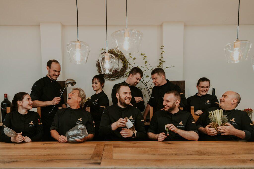 The group of chefs at Klosterhof Alpine Hideaway & Spa, dressed in black uniforms, stand and sit at a wooden table, smiling and laughing as they hold kitchen utensils and food in their hands under warm lighting.