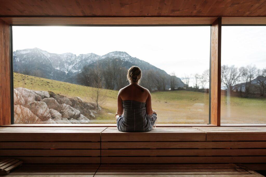 A woman sits relaxed on a wooden bench in the sauna and looks out of a large window with a great view of mountains and fields. She conveys a feeling of calm and contemplation.