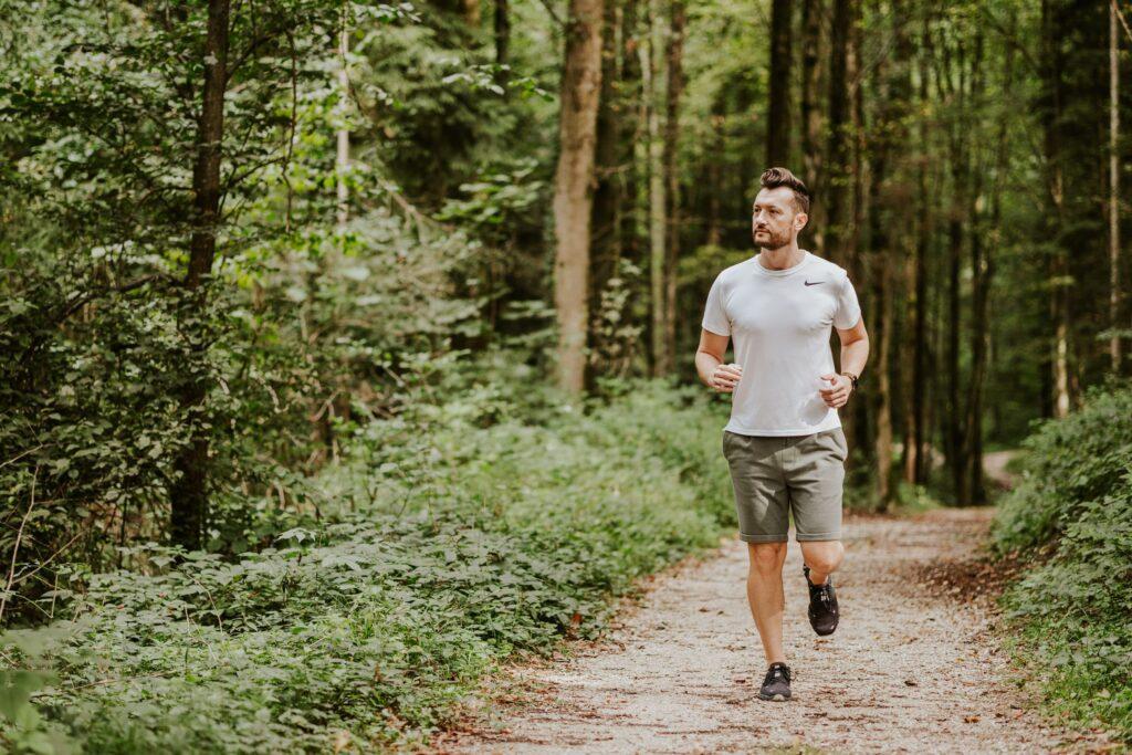A man jogs along a forest path, surrounded by lush green trees and foliage. He wears a white shirt and shorts and radiates a calm, focused energy.