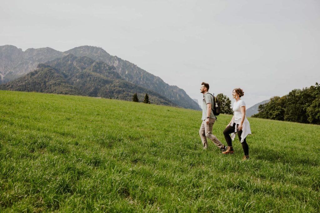 A man and a woman walk through a lush green meadow, surrounded by mountains and clear skies, conveying a sense of adventure and tranquility.