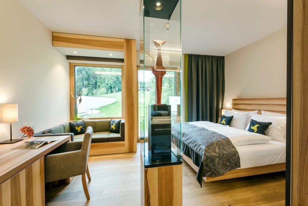 Modernes Hotelzimmer im Klosterhof Alpine Hideaway & Spa in Bayerisch Gmain bei Salzburg; mit Holzdeko, einem großen Bett, einer gemütlichen Sitzecke am Fenster mit toller Aussicht und einer Glassäule mit Kunstwerk.
