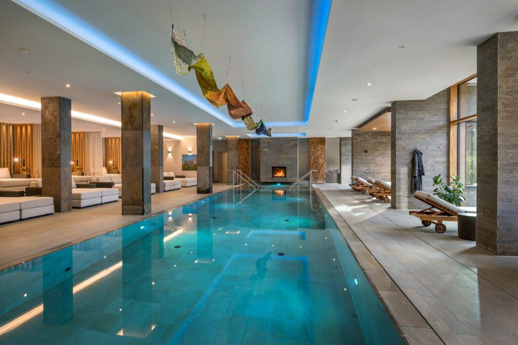The luxurious indoor pool at the Klosterhof Alpine Hideaway & Spa is surrounded by elegant lounge chairs and features a cozy fireplace. Artwork adorns the ceiling and creates a serene atmosphere.