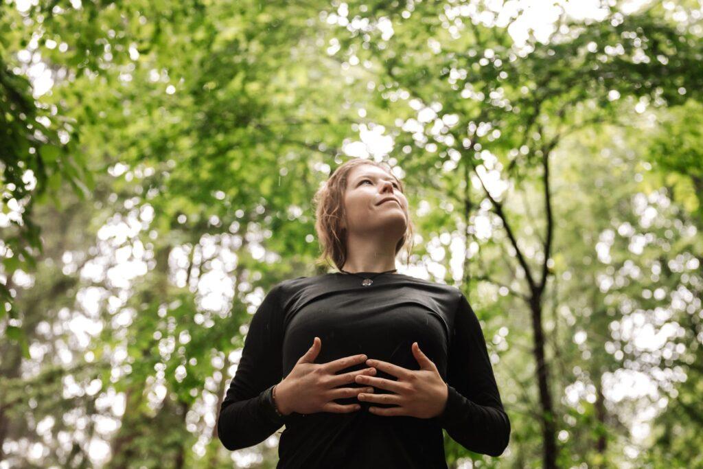 A woman in a black shirt stands in a lush green forest in the Berchtesgadener Land region, looking up with a calm expression and her hands on her chest. The scene conveys peace and a connection with nature.
