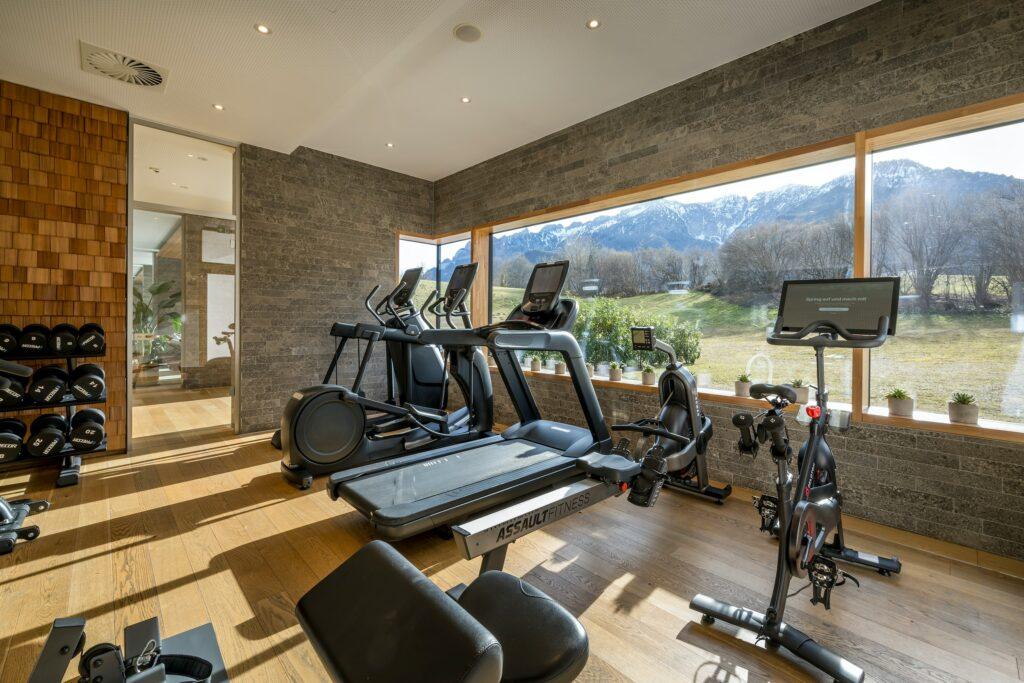 Modern fitness room in the sustainable Hotel Klosterhof in Germany with fitness equipment including treadmills and bikes, in front of large windows with a beautiful mountain view. Bright, inviting atmosphere.