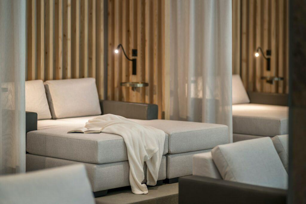 Comfortable lounge area with light gray chaise longues, covered with a soft beige blanket and an open book in the Klosterhof – Alpine Hideaway & Spa, a sustainable wellness hotel in Bavaria. The atmospheric lighting and the wooden paneling in the background create a calm atmosphere.