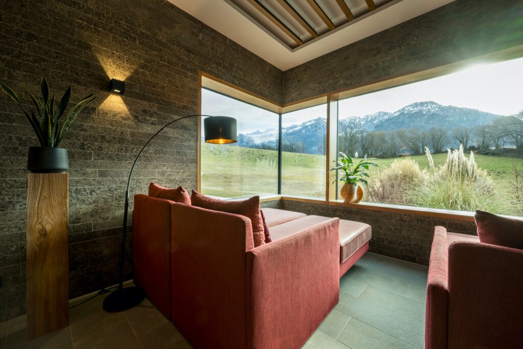 Modern relaxation room with red sofas and a black arched lamp, with large windows offering a peaceful view of green fields and snow-capped mountains at Klosterhof – Alpine Hideaway & Spa in Germany