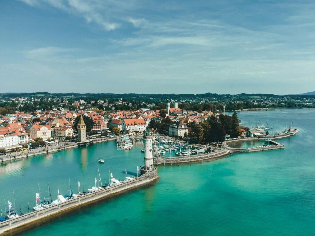Luftaufnahme der Küstenstadt Lindau am Bodensee mit ihren Gebäuden mit roten Dächern, einem Hafen mit Booten und einem Leuchtturm. Das Meer ist türkisblau und vermittelt eine ruhige und malerische Atmosphäre.