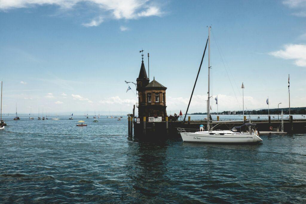 Historisches Hafengebäude am Bodensee mit einem Turm steht auf dem Wasser, umgeben von Segelbooten unter einem strahlend blauen Himmel. Ein weißes Segelboot fährt vorbei. Friedliche Atmosphäre.