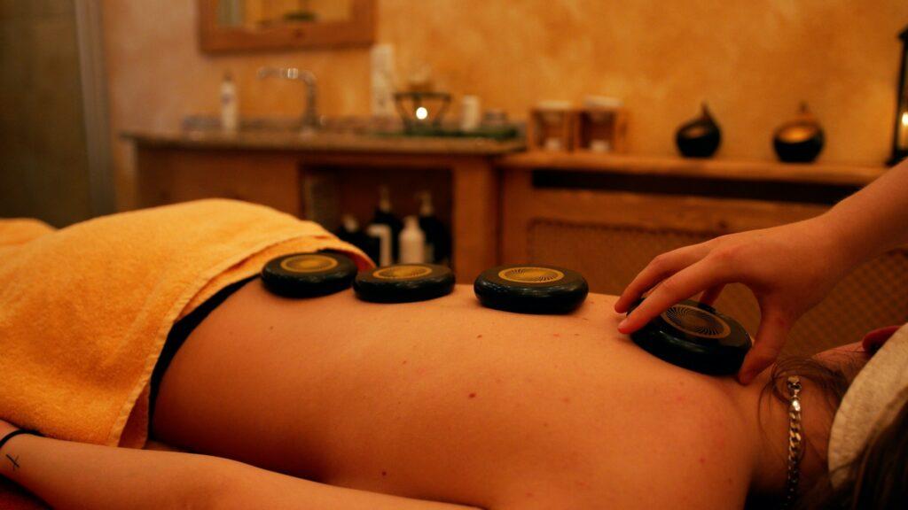 Eine Person, die in einem Spa eine Hot-Stone-Massage erhält. Vier schwarze Steine sind entlang des Rückens platziert, eine Hand richtet sie aus. Warme, entspannte Atmosphäre.
