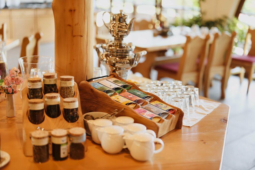 Eine Teestation am Bio-Frühstücksbuffet des Naturresorts Gerbehof mit verschiedenen Kräuterdosen, Teetassen, einem Samowar und einer bunten Teebox aus Holz auf einem Holztisch in einem gemütlichen Speisesaal.