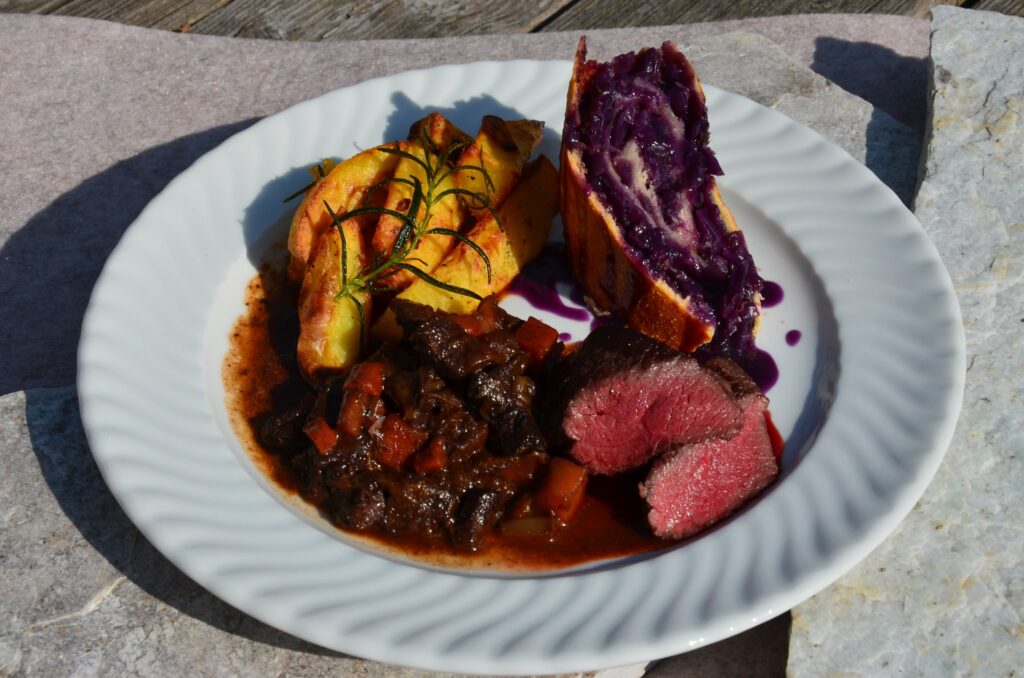 Ein weißer Teller mit Scheiben von medium-rare Bio-Fleisch, herzhaftem Eintopf, goldbraunen Kartoffelecken mit Rosmarin und leuchtend violettem Kohlkuchen. Das Gericht aus dem Naturresort Gerbehof ist farbenfroh und herzhaft.