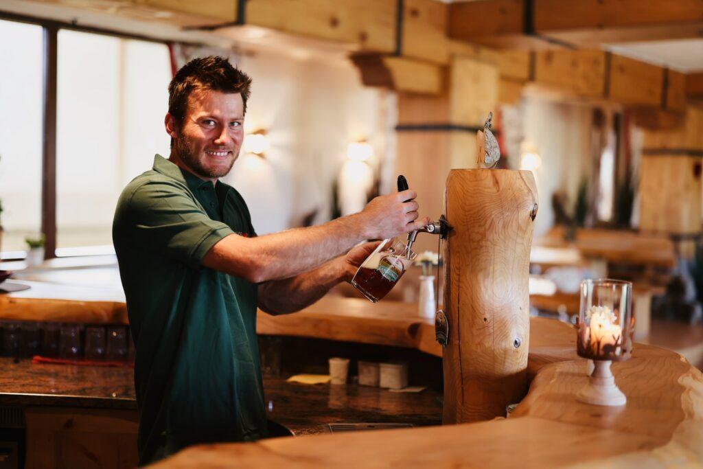 Ein lächelnder Mann in einem grünen Hemd zapft ein Pint Bier aus einem rustikalen Holzfass in der gemütlichen Bar des Bio-Hotels Naturresort Gerbehof mit Holzdekor und Kerzenlicht. Entspannte, einladende Atmosphäre.