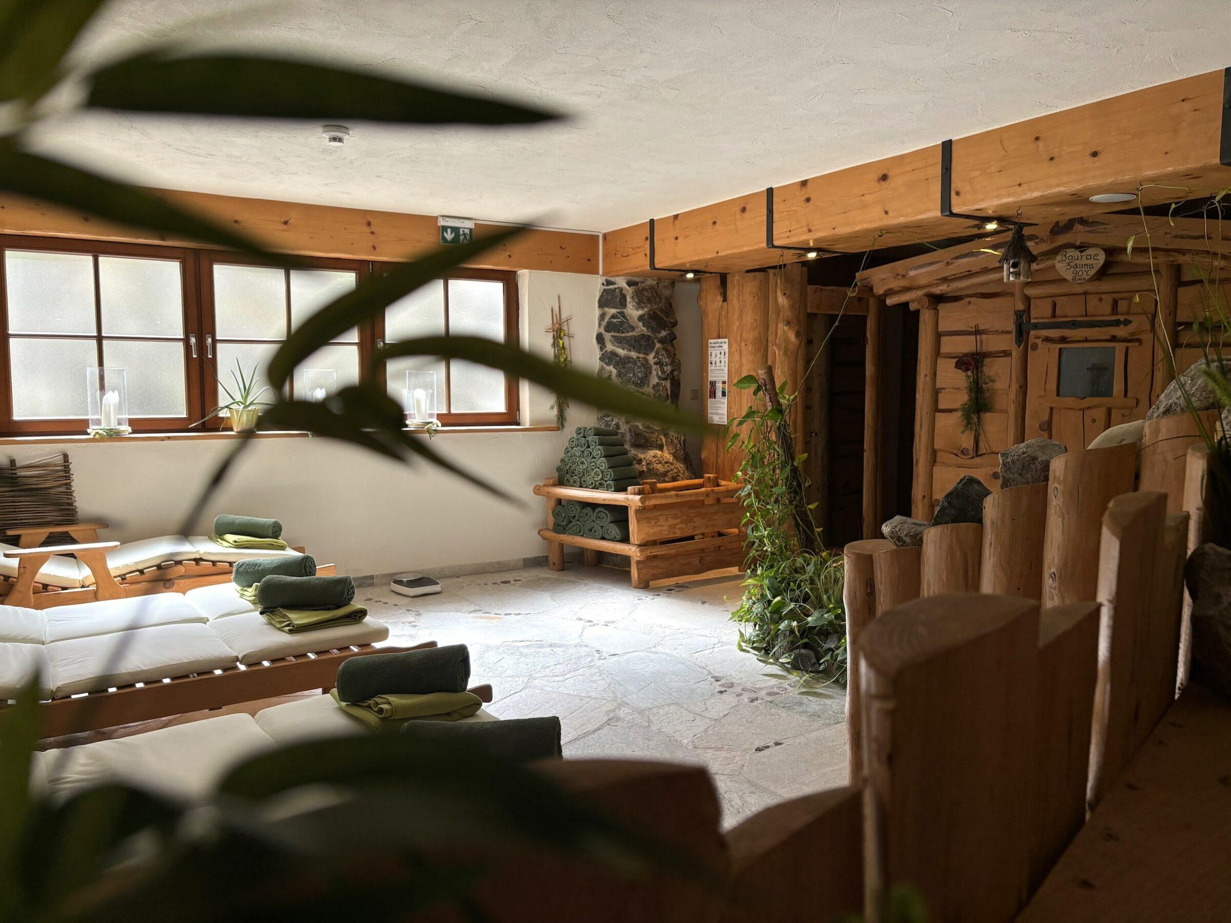 Gemütlicher Wellnessraum im Naturresort Gerbehof am Bodensee mit Holzliegen, die mit grünen Handtüchern bedeckt sind, Pflanzen, Akzenten aus Steinwänden und warmer, natürlicher Beleuchtung, die eine ruhige Atmosphäre schaffen.