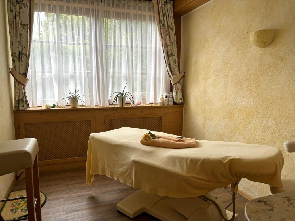 Beruhigender Massageraum mit einem Tisch, der mit einem weichen gelben Tuch bedeckt ist, auf dem ein Handtuch und eine Blume liegen. Sonnenlicht fällt durch die transparenten Vorhänge und beleuchtet die Pflanzen auf der Fensterbank.