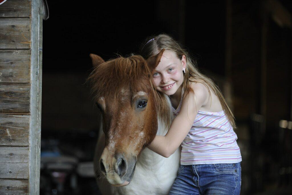 Ein junges Mädchen mit langen Haaren lächelt strahlend, während es ein braunes Pony in einem Stall umarmt. Die Szene strahlt Wärme und Freude aus, mit Holzwänden im Hintergrund.