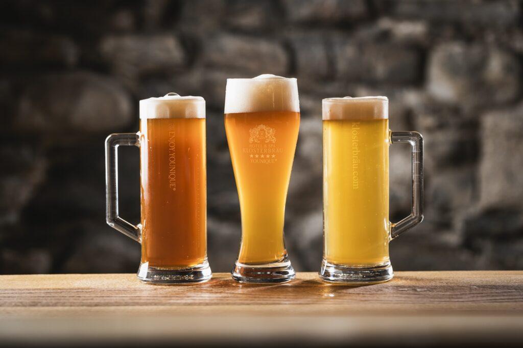 Three beer glasses in different shades from amber to yellow stand on a wooden table in front of a rustic stone wall. Each glass has a frothy head. The beer comes from the sustainable Hotel Klosterbräu's own brewery.