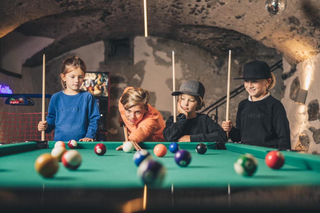 Four children stand around a pool table in the youth room of the sustainable Klosterbräu Hotel in Seefeld, concentrating and having fun. The room is cozy and rustically furnished, with stone walls and warm lighting.