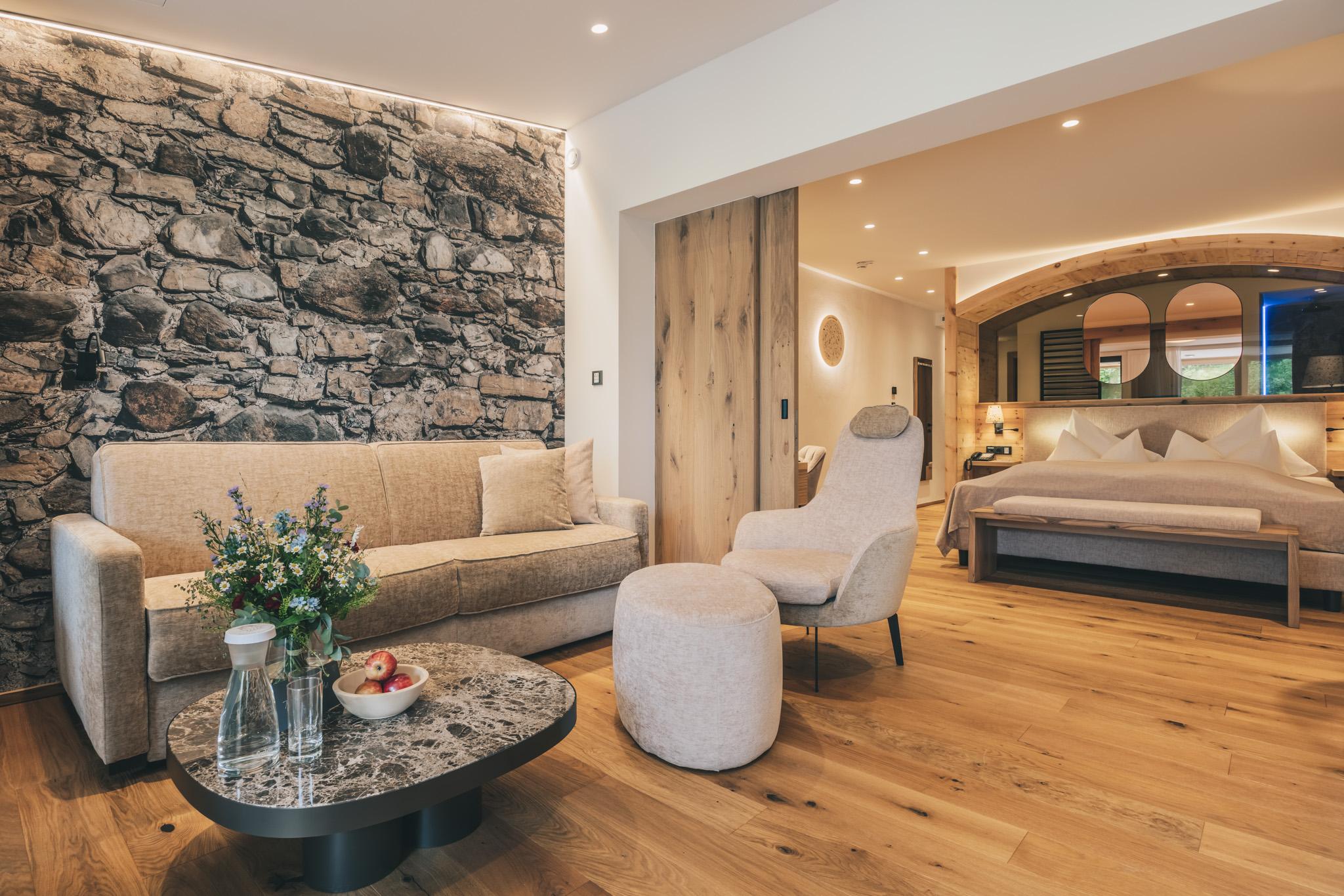 Modern hotel room made from natural materials in the sustainable Hotel Klosterbräu in Seefeld, Austria, with a cozy sofa, an accent wall made of stone, and elegant furnishings. A bed with soft lighting can be seen, creating a warm, inviting atmosphere.