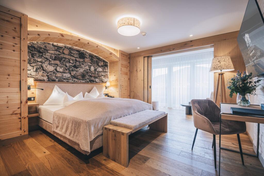 Cozy hotel room made from natural materials with rustic furnishings, an accent wall made of stone, wooden elements, a soft bed, a desk with chair, and soft lighting. From the sustainable Hotel Klosterbräu.