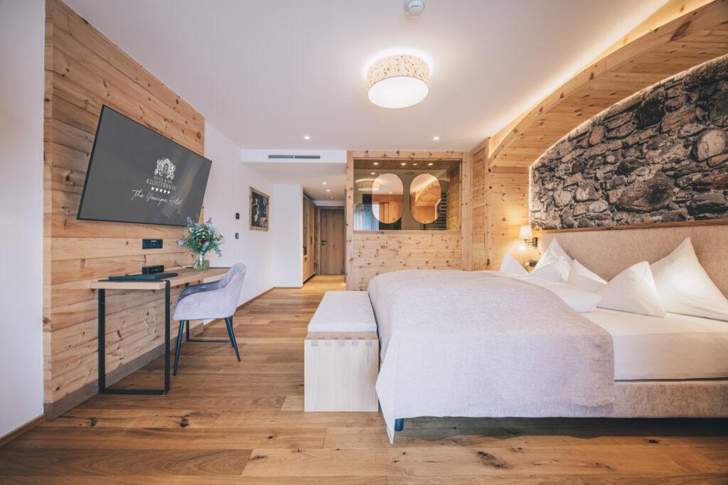 Modern hotel room with wood-paneled walls and floors, a comfortable double bed, large TV, desk, and soft lighting at the Austrian wellness hotel Klosterbräu. Warm, inviting, and elegant atmosphere.