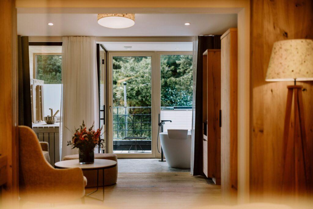 A cozy, elegant room with a large window overlooking greenery, equipped with a freestanding bathtub, an armchair, a round table with flowers, and warm lighting.