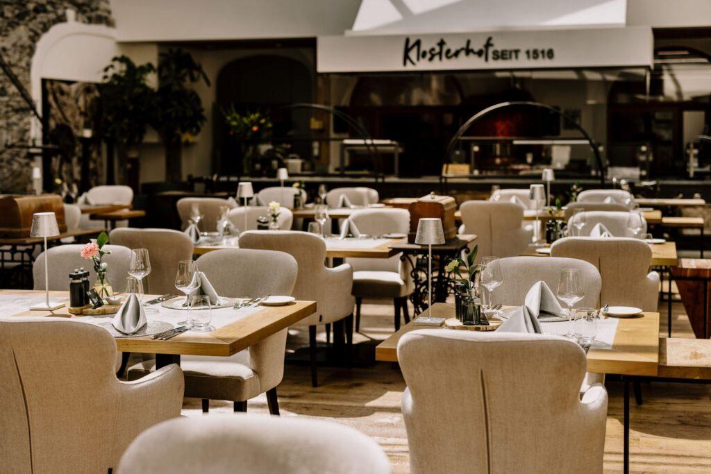 Elegant restaurant interior in the sustainable Hotel Klosterbräu in Austria with beige chairs and wooden tables set with glasses and napkins. Soft lighting and a cozy, inviting atmosphere.