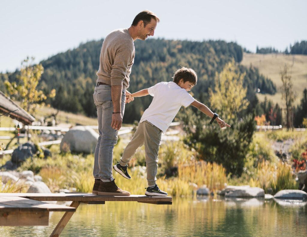 A man and a child on a wooden platform above a pond, surrounded by the picturesque Austrian landscape around Seefeld with green hills and trees, share a joyful moment and point forward.